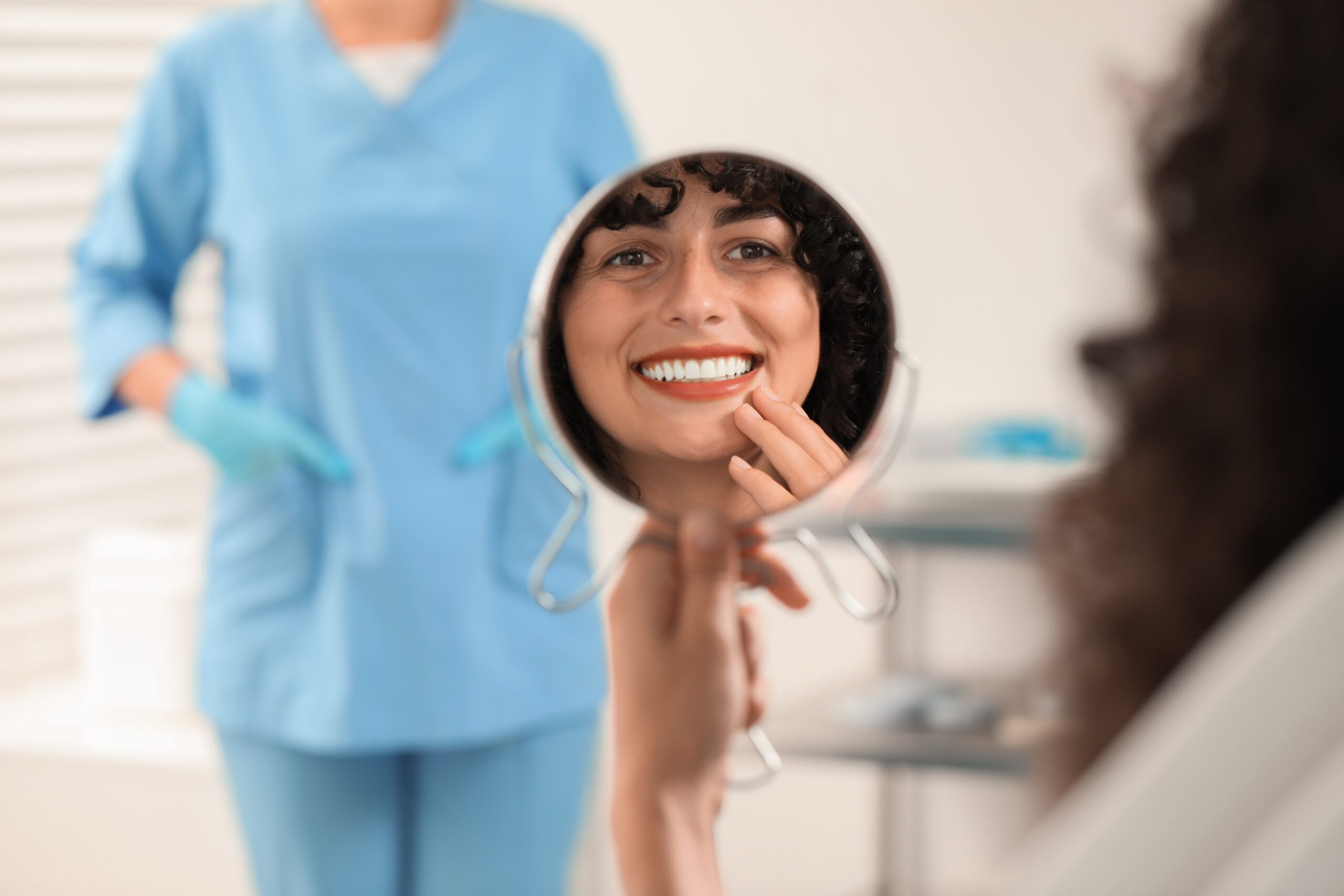 young woman smiling in mirror, doctor in background, happy with new cosmetic bonding